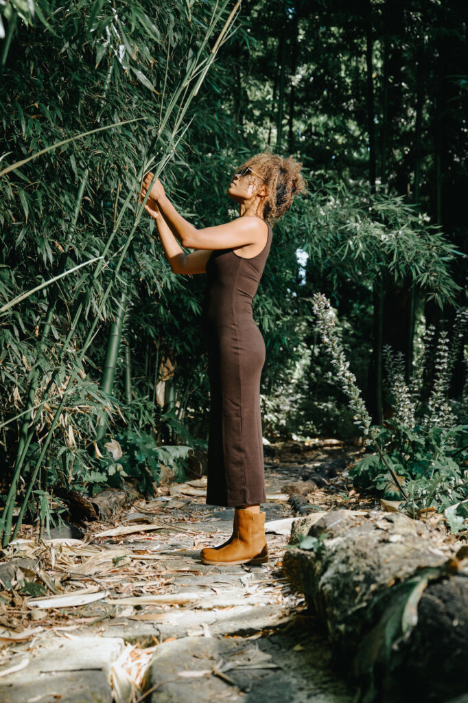 Woman in a brown sleeveless dress and camel boots standing on a forest path, interacting with bamboo.