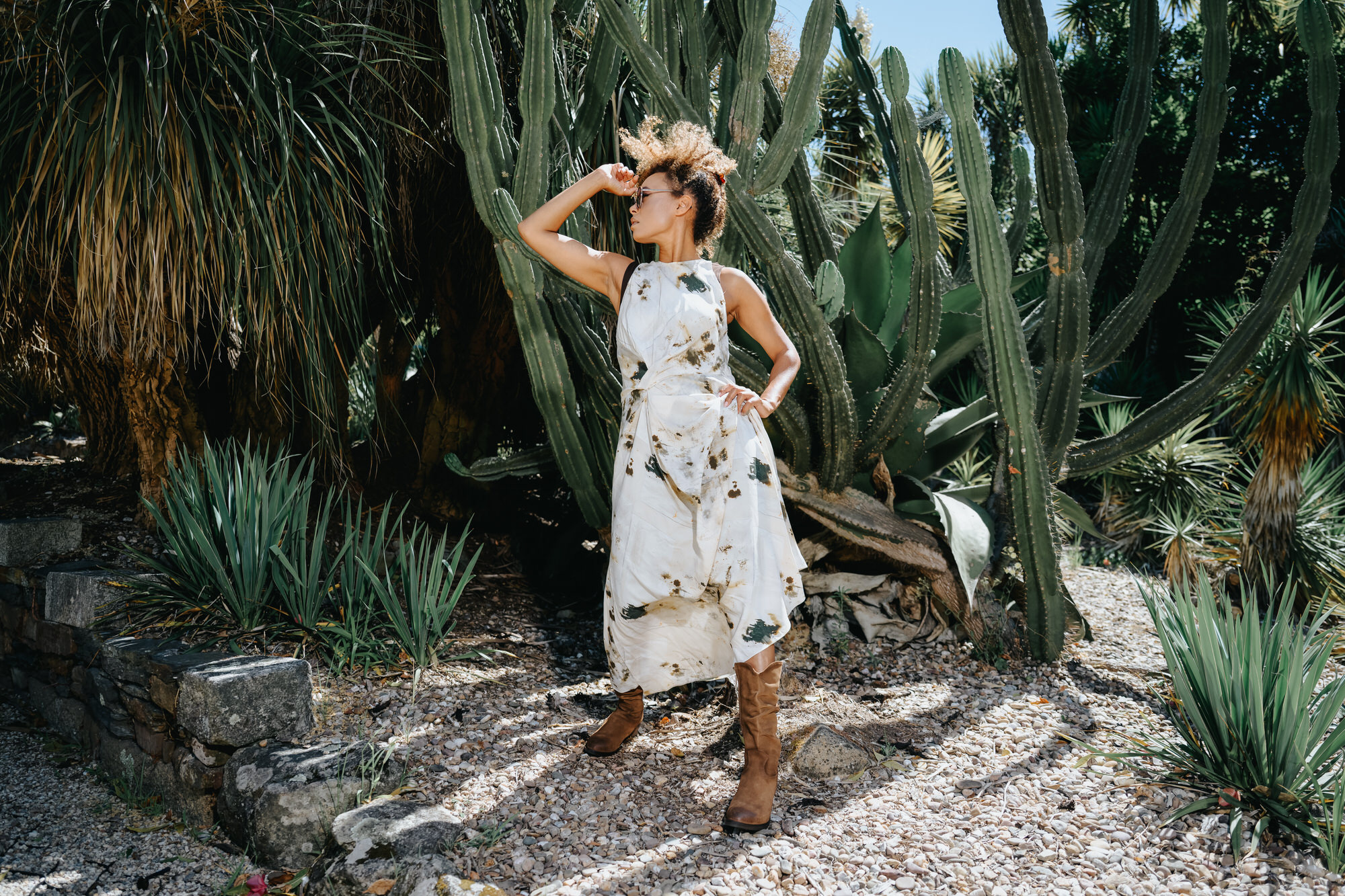 Woman posing confidently in front of large cacti, wearing a white patterned dress and brown boots.