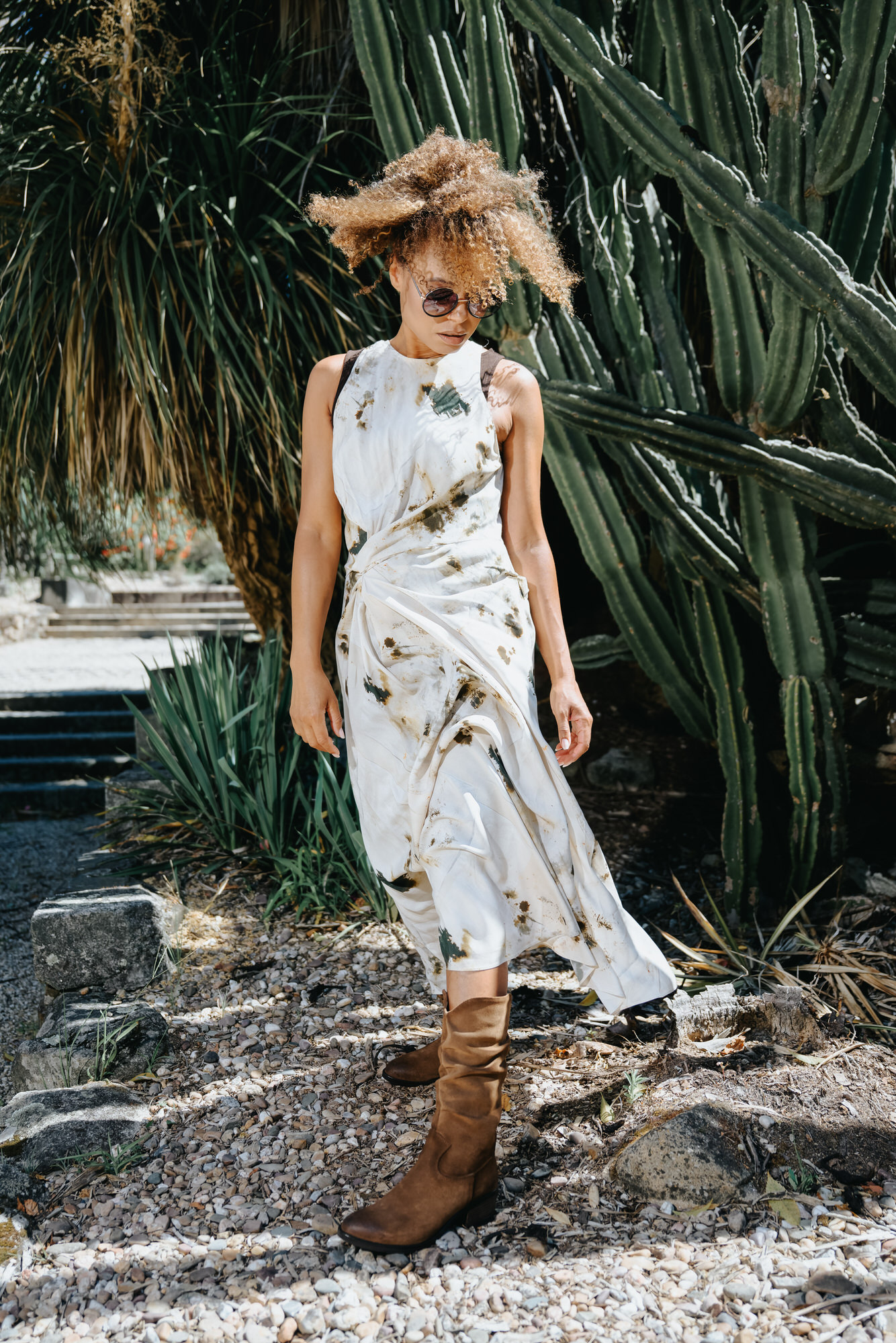 Woman in a flowing white dress with earth-toned patterns, wearing slouchy brown boots and sunglasses, standing in front of tall cacti on a rocky path.