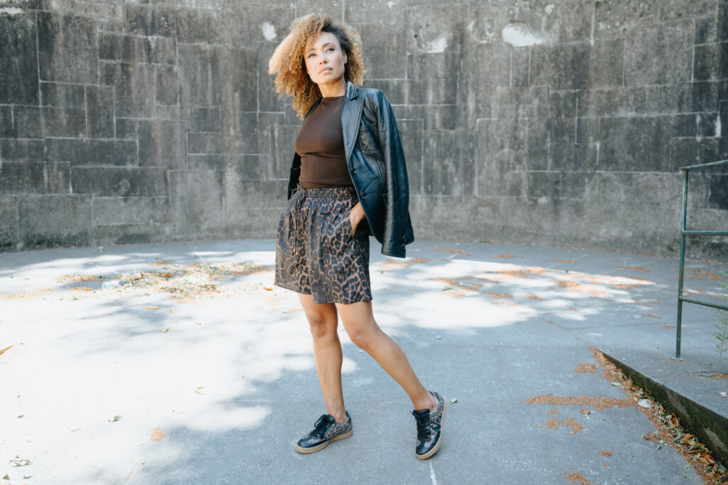 Woman standing confidently on a sunlit concrete surface, wearing a black leather jacket, brown top, leopard print shorts, and matching sneakers.