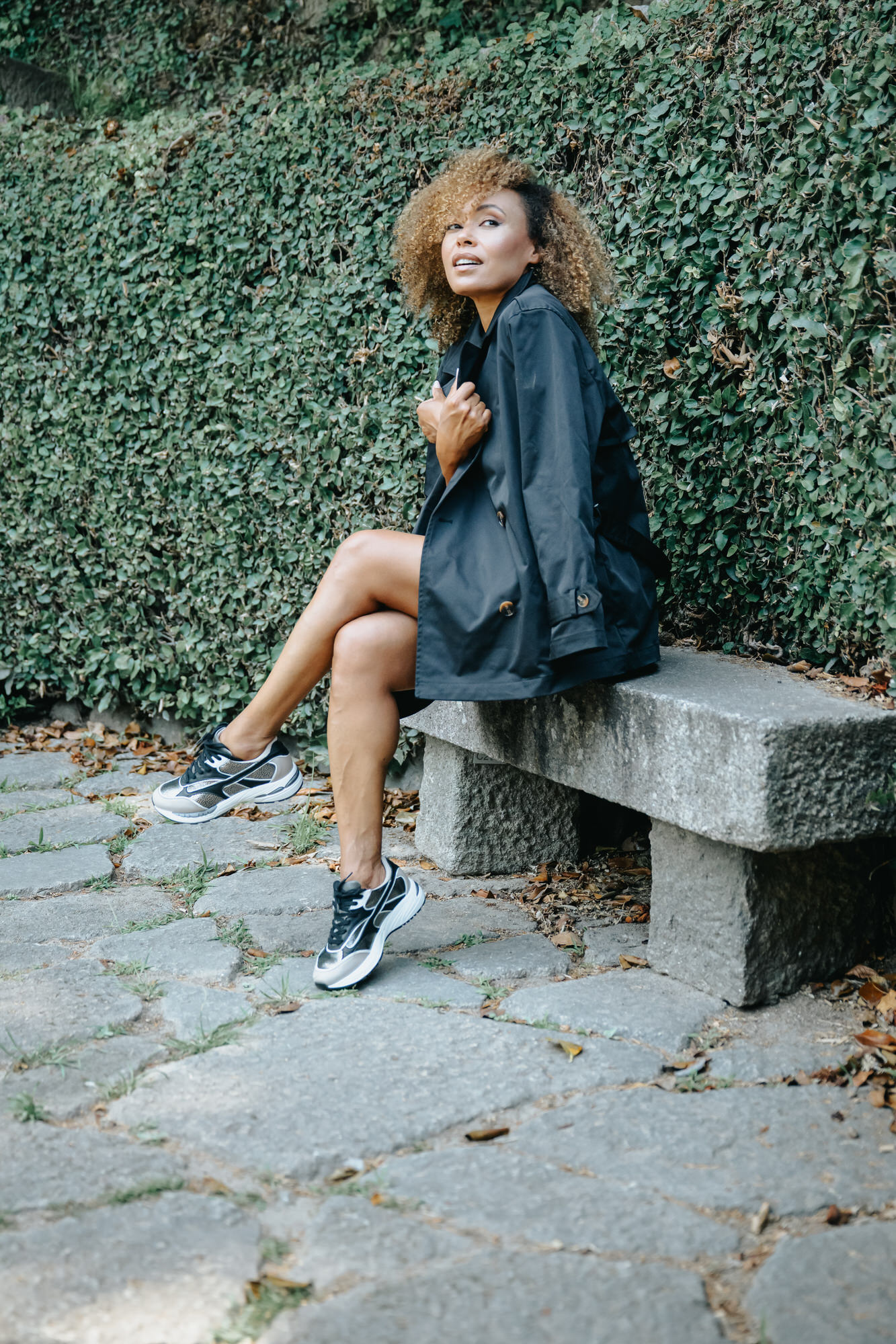 Woman sitting on a stone bench, wearing black-and-white sneakers and a black trench coat, with a leafy green hedge behind her.