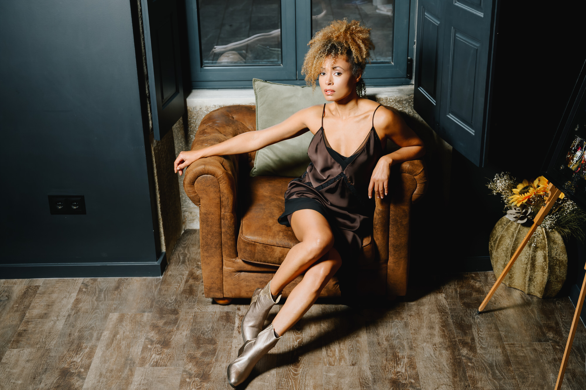 Woman sitting in a vintage leather armchair wearing AQA metallic bronze pointed-toe ankle boots and a dark slip dress.