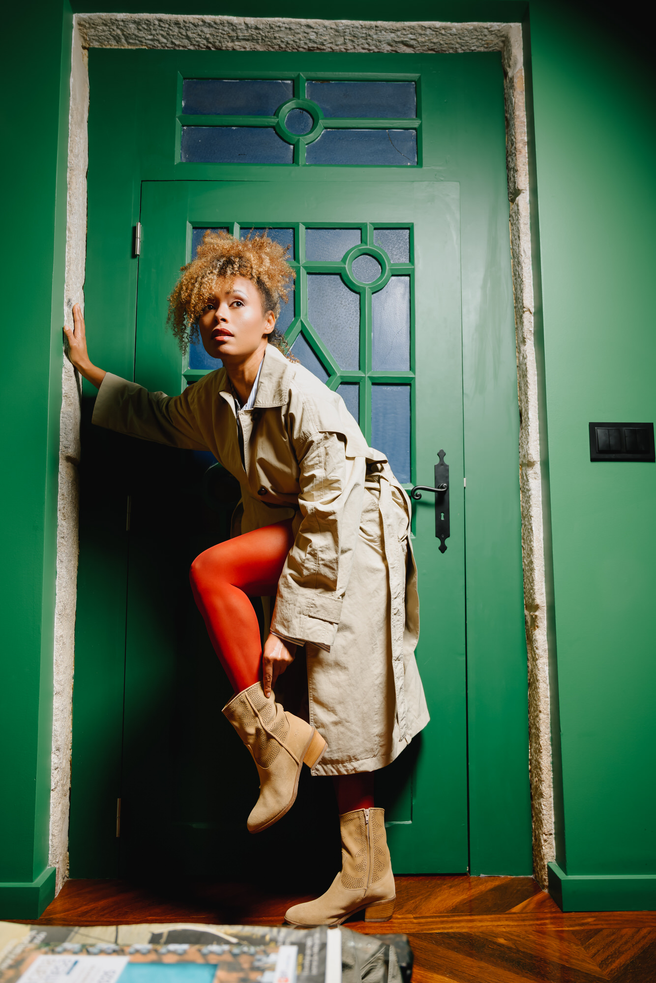 A person wearing AQA beige suede ankle boots with decorative laser-cut perforations and a stacked block heel. The boots are styled with vibrant orange tights and a trench coat, showcasing the AQA footwear seasonal collection.