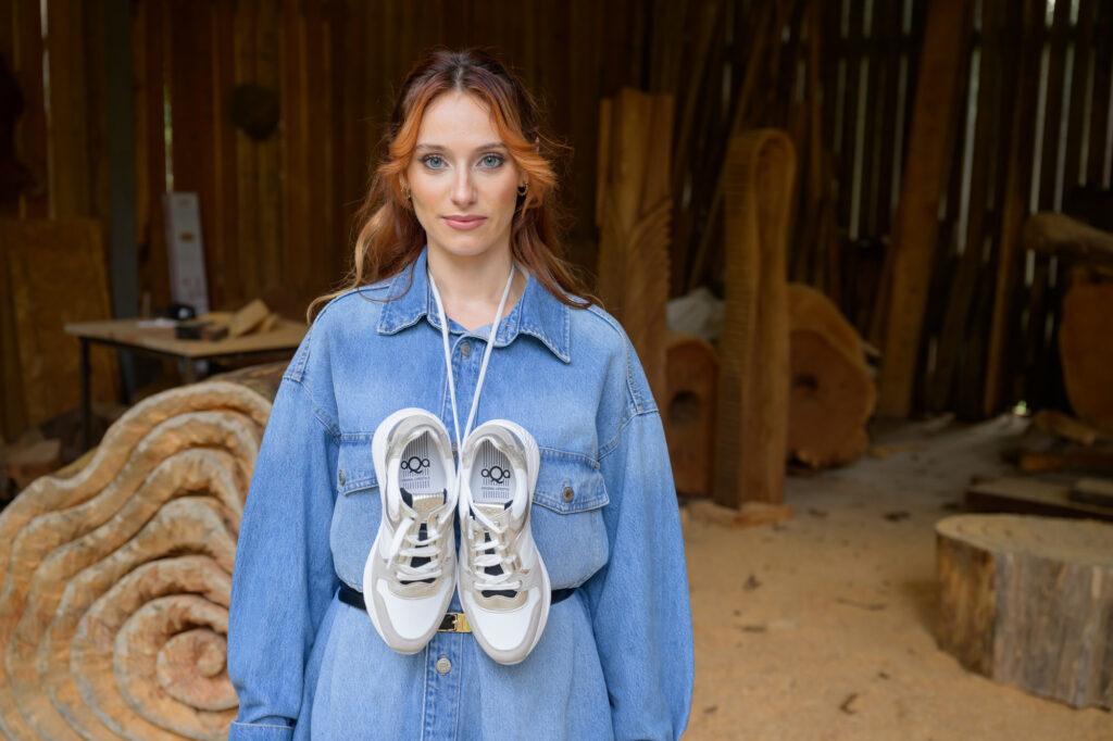 Woman wearing a denim dress with AQA white and gold sneakers hanging around her neck in a rustic wood workshop - Spring/Summer 2026.