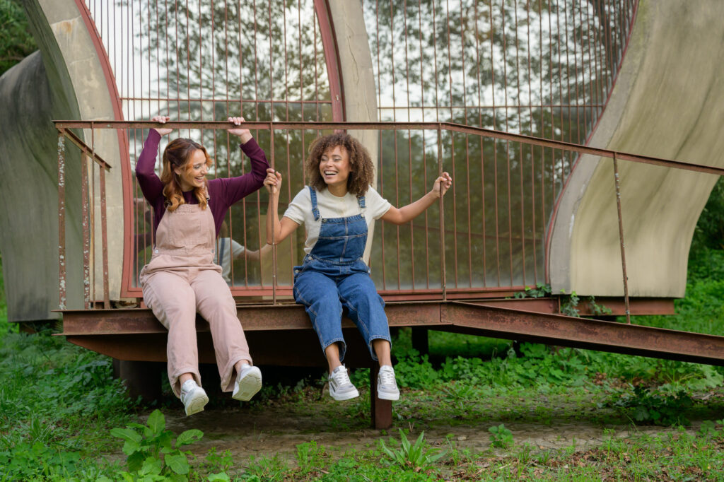 Two women sitting on steps, wearing matching navy trousers and AQA metallic silver chunky sneakers - Spring/Summer 2026 Collection.