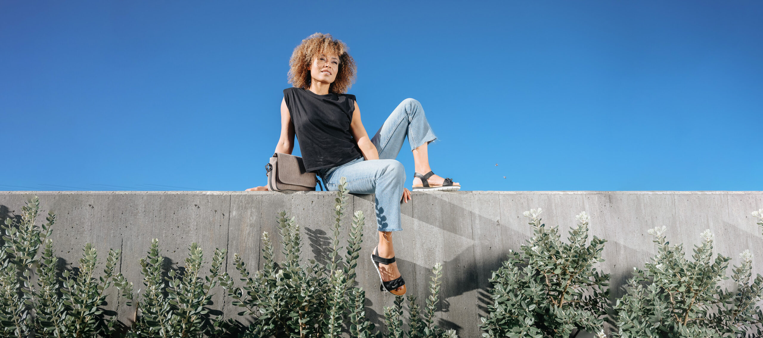 A woman with curly hair sits on a concrete ledge wearing light-wash jeans and a black sleeveless top, showcasing AQA Shoes black casual sport sandals against a clear blue sky.