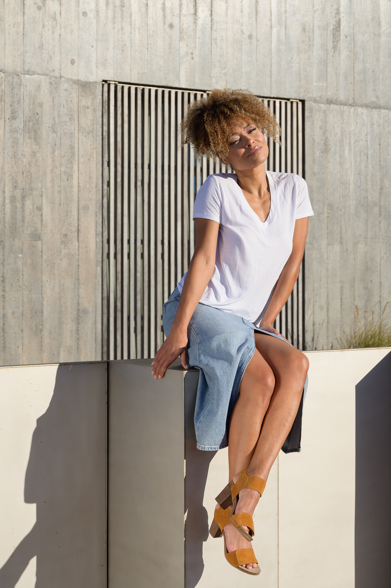 Woman sitting on a concrete wall wearing a white t-shirt, denim skirt, and AQA Shoes tan suede heeled sandals with ankle straps under bright sunlight.